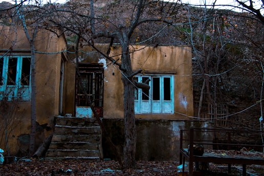 An abandoned house stands amid barren trees. The structure appears weathered, with peeling paint and broken windows. The steps leading to the entrance are cracked, and the surrounding ground is covered with dry leaves. The atmosphere is desolate and haunting.