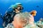 A scuba diver is underwater, surrounded by vibrant marine life. The diver is wearing a black wetsuit and mask, swimming alongside orange and white fish near a large pink sea anemone nestled in a coral reef. The ocean water is a deep blue, and additional small fish are visible in the background.