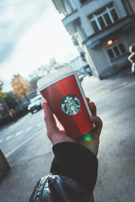 A hand holding a to-go cup with the Sugar and Spice Coffee logo against a backdrop of autumn leaves.