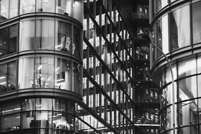 A modern architectural structure featuring curved glass facades with visible office interiors. The reflections and lighting create a complex pattern of lines and shapes, showcasing the sleek design of a contemporary high-rise building.