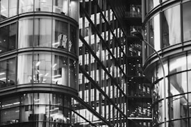 A modern architectural structure featuring curved glass facades with visible office interiors. The reflections and lighting create a complex pattern of lines and shapes, showcasing the sleek design of a contemporary high-rise building.