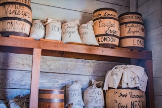 Wooden shelves are stocked with barrels and cloth bags. The barrels are labeled with product names like 'Vinegar' and 'Crushed Sugar' and indicate their origin as London. The cloth bags are labeled with items such as 'Rice' and 'Fine Flour'. The setting appears to resemble an old-fashioned pantry or storage room with a rustic and vintage feel.