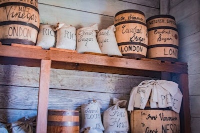 Wooden shelves are stocked with barrels and cloth bags. The barrels are labeled with product names like 'Vinegar' and 'Crushed Sugar' and indicate their origin as London. The cloth bags are labeled with items such as 'Rice' and 'Fine Flour'. The setting appears to resemble an old-fashioned pantry or storage room with a rustic and vintage feel.