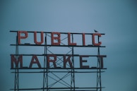Red neon letters spell out 'PUBLIC MARKET' against a cloudy blue sky. A bird, possibly a seagull, is perched on the structure.