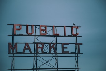 Red neon letters spell out 'PUBLIC MARKET' against a cloudy blue sky. A bird, possibly a seagull, is perched on the structure.