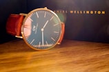 A sophisticated wristwatch with a minimalist design featuring a large round face and simple hour markers, complemented by a rich brown leather strap. The watch is set on a wooden surface with a blurred dark background, hinting at a luxury brand's packaging.