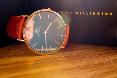 A sophisticated wristwatch with a minimalist design featuring a large round face and simple hour markers, complemented by a rich brown leather strap. The watch is set on a wooden surface with a blurred dark background, hinting at a luxury brand's packaging.