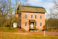 A two-story red brick building with multiple tall windows, situated in a wooded area with bare trees. The building features a chimney and has a wooden railing along its small porch. The ground around the building is covered in fallen leaves, suggesting an autumn or winter setting.