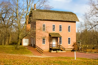 A two-story red brick building with multiple tall windows, situated in a wooded area with bare trees. The building features a chimney and has a wooden railing along its small porch. The ground around the building is covered in fallen leaves, suggesting an autumn or winter setting.