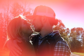 A couple embraces and smiles at each other. Sunlight creates a warm, red-orange glow around them, enhancing the romantic atmosphere. They are casually dressed, standing in an outdoor setting with a backdrop of trees under a clear sky.