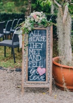 A decorative chalkboard sign displays the phrase 'ALL BECAUSE THEY MET AND FELL in love' with a pink heart drawn near the bottom. The sign is adorned with a bouquet of white, pink, and purple flowers, along with green foliage, positioned at the top. It's placed on a gravel path next to a large terracotta plant pot and a tree draped with Spanish moss. In the background, there are several black woven chairs set up outdoors, surrounded by greenery.