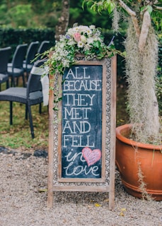 A decorative chalkboard sign displays the phrase 'ALL BECAUSE THEY MET AND FELL in love' with a pink heart drawn near the bottom. The sign is adorned with a bouquet of white, pink, and purple flowers, along with green foliage, positioned at the top. It's placed on a gravel path next to a large terracotta plant pot and a tree draped with Spanish moss. In the background, there are several black woven chairs set up outdoors, surrounded by greenery.