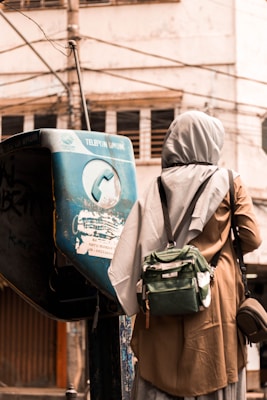 A person wearing a light gray headscarf and brown clothing stands in front of an old public telephone. Wires crisscross the background with a worn, urban environment visible.