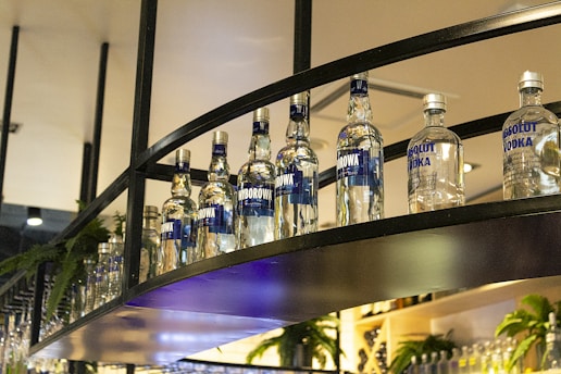 A row of clear glass vodka bottles displayed on a curved metal shelf, with ferns and soft lighting in the background creating an elegant bar setting.