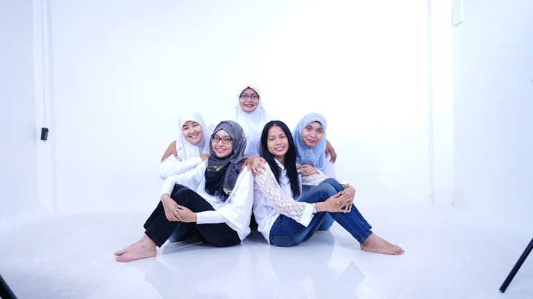 Five women are sitting together on the floor in a bright, minimalistic setting. They are wearing white tops and jeans, with some wearing headscarves. They appear happy and relaxed, with some of them embracing each other.