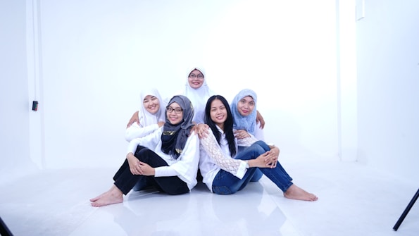 Five women are sitting together on the floor in a bright, minimalistic setting. They are wearing white tops and jeans, with some wearing headscarves. They appear happy and relaxed, with some of them embracing each other.