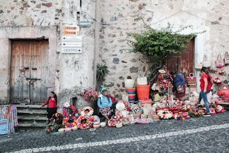 people sitting and standing by the brick wall beside roadway with assorted display products on the ground