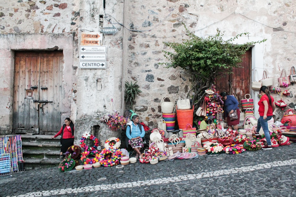 Personas sentadas y de pie junto a la pared de ladrillos al lado de la carretera con una variedad de productos de exhibición en el suelo