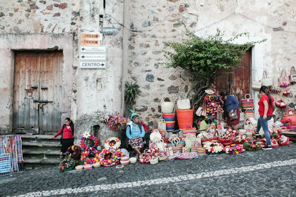 People sitting and standing by the brick wall beside roadway with assorted display products on the ground — Taxco, Mexico