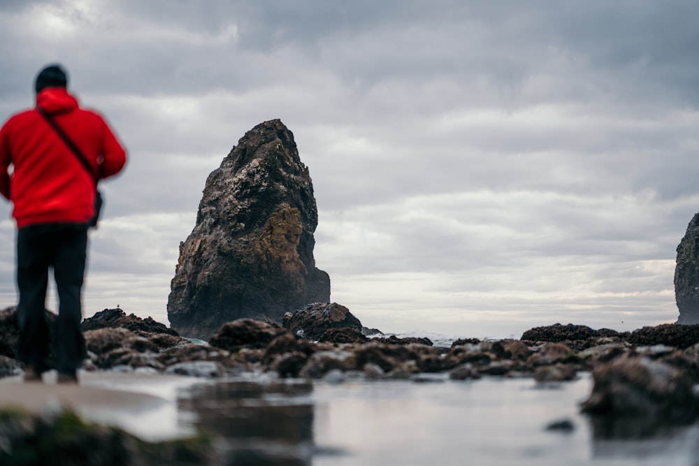 man in red hoodie standing on rocky shore under cloudy sky during daytime