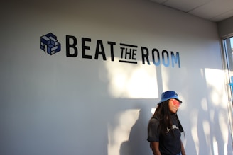A person wearing a blue hat and sunglasses stands in front of a wall with the words 'Beat The Room' and a cube logo. Sunlight casts shadows on the wall, and a window is visible on the right side.
