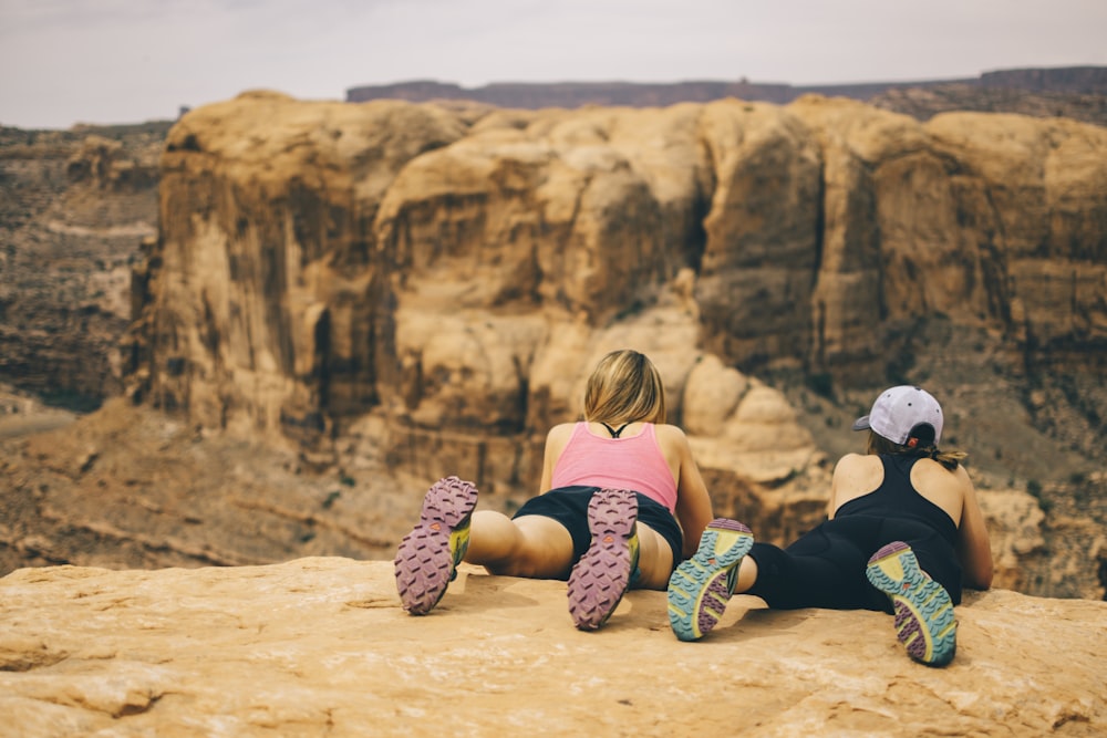 two woman lying face down on top of rock cliff overlooking cliff at daytime