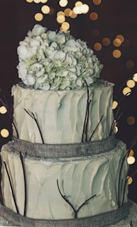 A three-tier cake with a rustic design, featuring smooth white frosting and decorated with a large cluster of white flowers on top. The tiers are wrapped with a textured gray ribbon, and thin branches are placed around each layer. The background is softly lit with warm, blurred lights, adding a calm ambiance.
