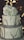 A three-tier cake with a rustic design, featuring smooth white frosting and decorated with a large cluster of white flowers on top. The tiers are wrapped with a textured gray ribbon, and thin branches are placed around each layer. The background is softly lit with warm, blurred lights, adding a calm ambiance.