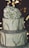 A three-tier cake with a rustic design, featuring smooth white frosting and decorated with a large cluster of white flowers on top. The tiers are wrapped with a textured gray ribbon, and thin branches are placed around each layer. The background is softly lit with warm, blurred lights, adding a calm ambiance.