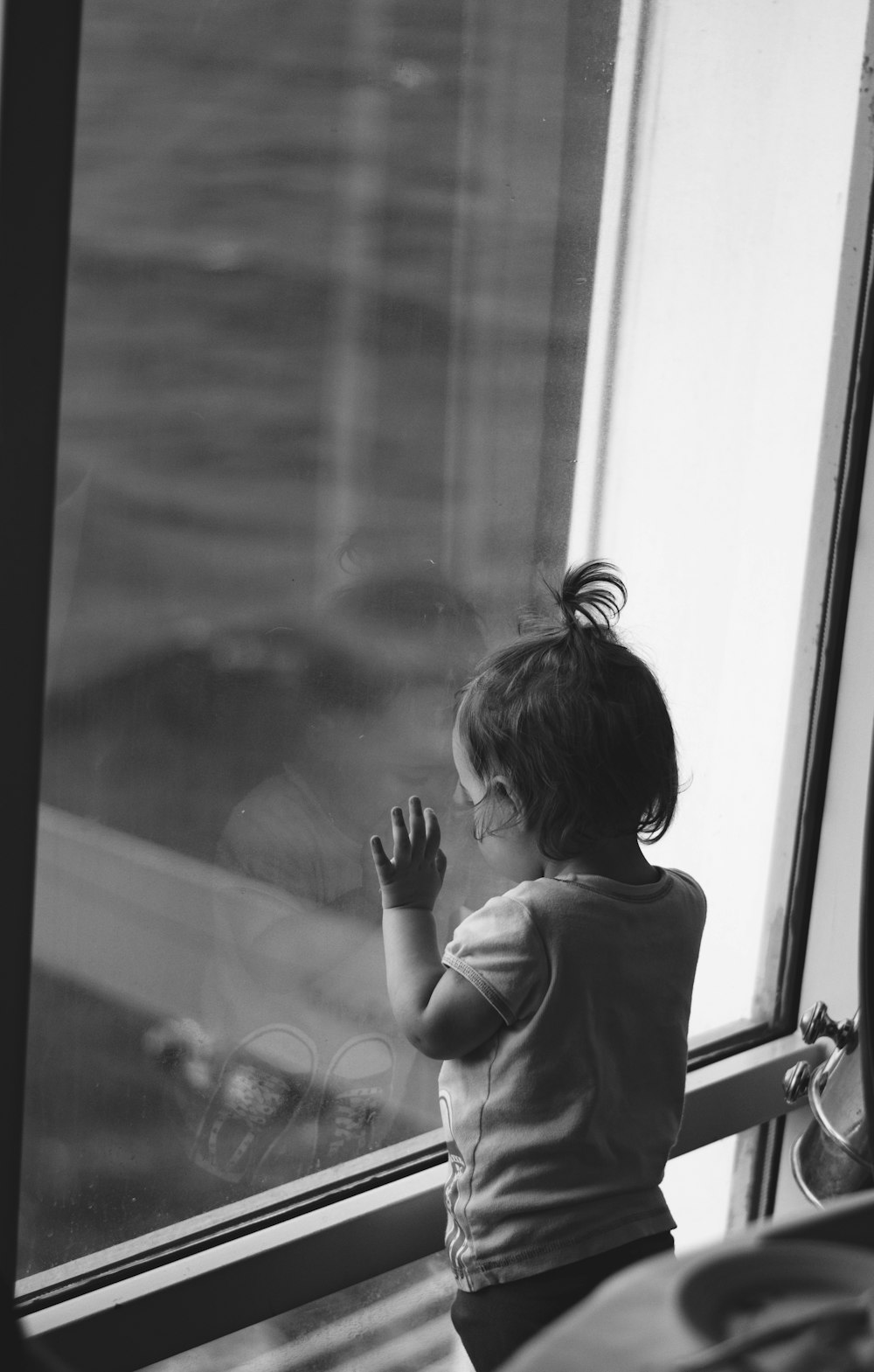 baby leaning on glass window while looking down in greyscale photography