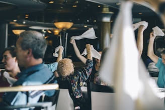 People are sitting in a restaurant or dining area, some of them raising white napkins in the air. The setting includes hanging lights and tables, with a focus on social interaction and enjoyment.