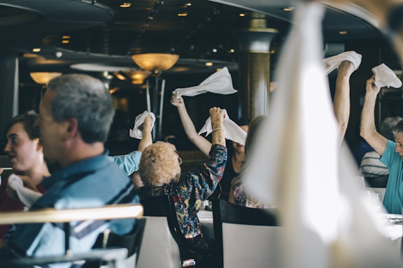 People are sitting in a restaurant or dining area, some of them raising white napkins in the air. The setting includes hanging lights and tables, with a focus on social interaction and enjoyment.