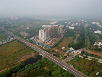 An aerial view of a developing urban area featuring several high-rise buildings under construction. The surroundings include a network of roads, greenery, and scattered residential and commercial structures.