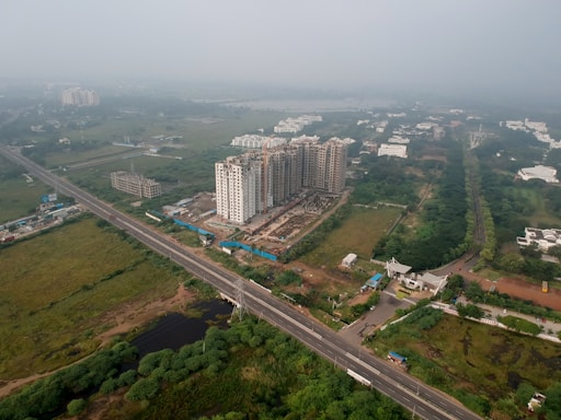 An aerial view of a developing urban area featuring several high-rise buildings under construction. The surroundings include a network of roads, greenery, and scattered residential and commercial structures.