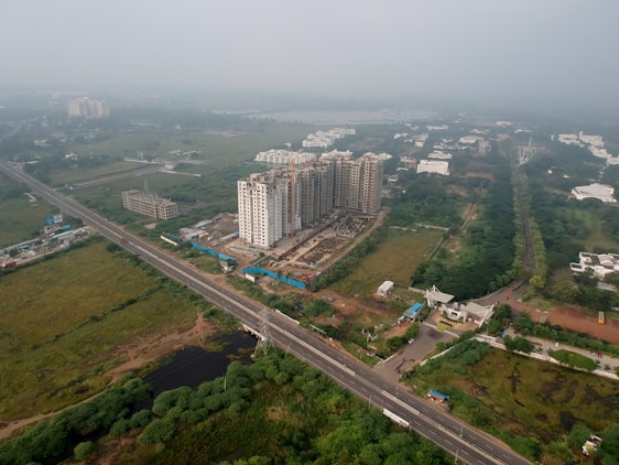 An aerial view of a developing urban area featuring several high-rise buildings under construction. The surroundings include a network of roads, greenery, and scattered residential and commercial structures.