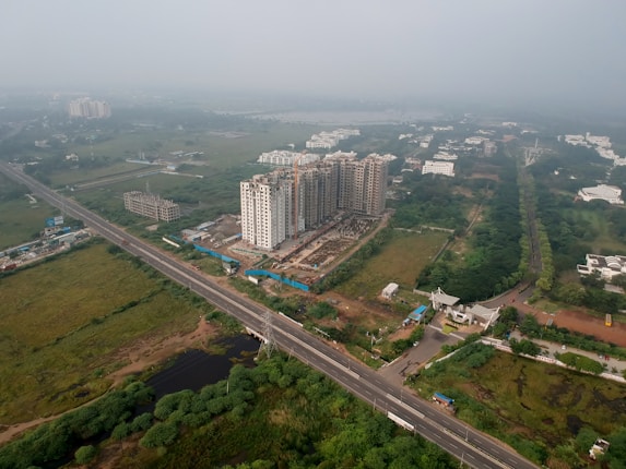 An aerial view of a developing urban area featuring several high-rise buildings under construction. The surroundings include a network of roads, greenery, and scattered residential and commercial structures.