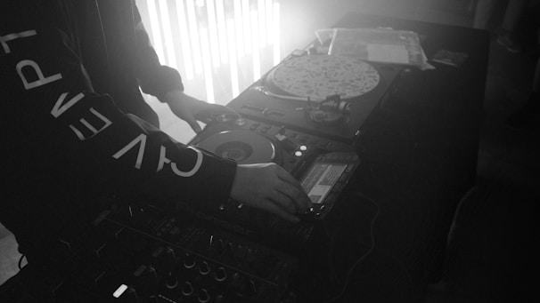 A person is operating DJ equipment with a focus on a turntable and mixer. The setting appears to be dimly lit, possibly a club or party, with light strips adding to the atmosphere. The person's clothing is adorned with large printed letters.