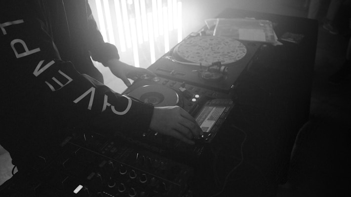 A person is operating DJ equipment with a focus on a turntable and mixer. The setting appears to be dimly lit, possibly a club or party, with light strips adding to the atmosphere. The person's clothing is adorned with large printed letters.