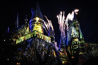 A majestic castle illuminated at night with vibrant, colorful lights and fireworks exploding in the sky above. The architecture features tall spires and intricate designs, creating a magical and enchanting atmosphere.