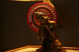 A bronze-colored statue of a serene, meditating figure is depicted with a peaceful expression, seated against a vibrant, intricately decorated red halo with metallic beads. The lighting casts a warm glow, highlighting the statue's smooth contours.