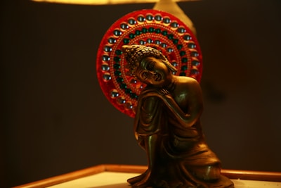 A bronze-colored statue of a serene, meditating figure is depicted with a peaceful expression, seated against a vibrant, intricately decorated red halo with metallic beads. The lighting casts a warm glow, highlighting the statue's smooth contours.