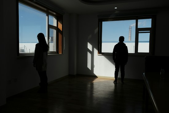 Two silhouetted figures stand near large windows with city views outside. The room is dimly lit with sunlight streaming in through the windows, casting shadows on the wooden floor. One person is near the left window, and the other is near the right window, facing outside.