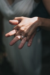 Two hands are interlocked in an intimate gesture, focusing on a woman's hand adorned with an engagement ring. Soft lighting accentuates the tenderness and warmth of the moment.