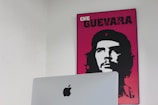 A minimalist room features a bright poster on the wall, depicting a stylized black and white portrait of Che Guevara with a vibrant pink background. In the foreground, there is an Apple computer with a prominent logo visible.