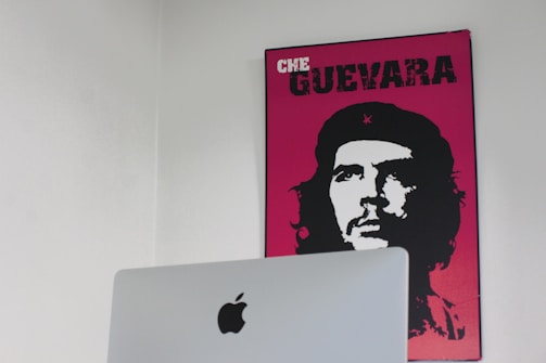 A minimalist room features a bright poster on the wall, depicting a stylized black and white portrait of Che Guevara with a vibrant pink background. In the foreground, there is an Apple computer with a prominent logo visible.