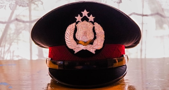 A formal dark hat featuring an intricate badge design with laurels and stars, resting on a reflective wooden surface. The light source from the back creates a silhouette effect, highlighting the hat's contours.