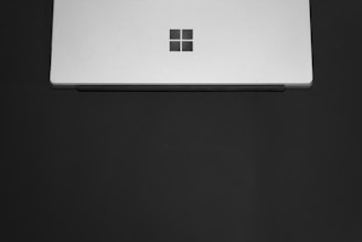 A sleek, silver laptop partially closed with a minimalist logo on the center of the lid. The laptop is set against a plain dark background, emphasizing its modern design.