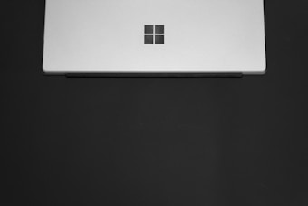 A sleek, silver laptop partially closed with a minimalist logo on the center of the lid. The laptop is set against a plain dark background, emphasizing its modern design.