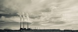 A sepia-toned photograph features an industrial scene with three tall smokestacks emitting smoke into a cloudy sky. The landscape includes power lines and a barren, rocky foreground, creating a stark and expansive view.