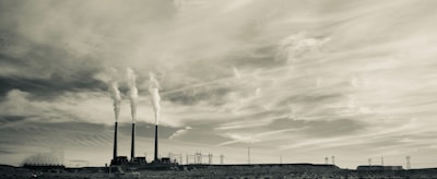 A sepia-toned photograph features an industrial scene with three tall smokestacks emitting smoke into a cloudy sky. The landscape includes power lines and a barren, rocky foreground, creating a stark and expansive view.